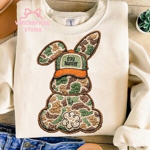 May include: Cream-colored sweatshirt with an embroidered Easter bunny design. The bunny is wearing a camouflage hat with the text "egg hunter." The bunny's body is also in a camouflage pattern, with a flower detail at the bottom.