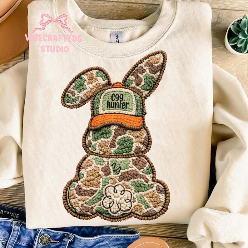 May include: Cream-colored sweatshirt with an embroidered Easter bunny design. The bunny is wearing a camouflage hat with the text "egg hunter." The bunny's body is also in a camouflage pattern, with a flower detail at the bottom.