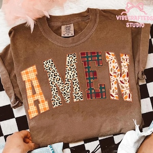 Può includere: Una maglietta marrone Comfort Colors con la scritta "AMEN" in grandi lettere decorative. La lettera "A" è a quadri arancioni, "M" è un motivo leopardato, "E" è a quadri verdi e rossi e "N" è con foglie autunnali. La maglietta è piegata.
