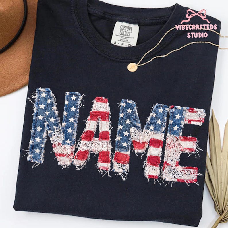 May include: Dark blue t-shirt with the word "NAME" appliqued in a patriotic design. The letters feature a distressed American flag pattern with red and white stripes and blue star-studded sections. A gold necklace and a Comfort Colors tag are also visible.