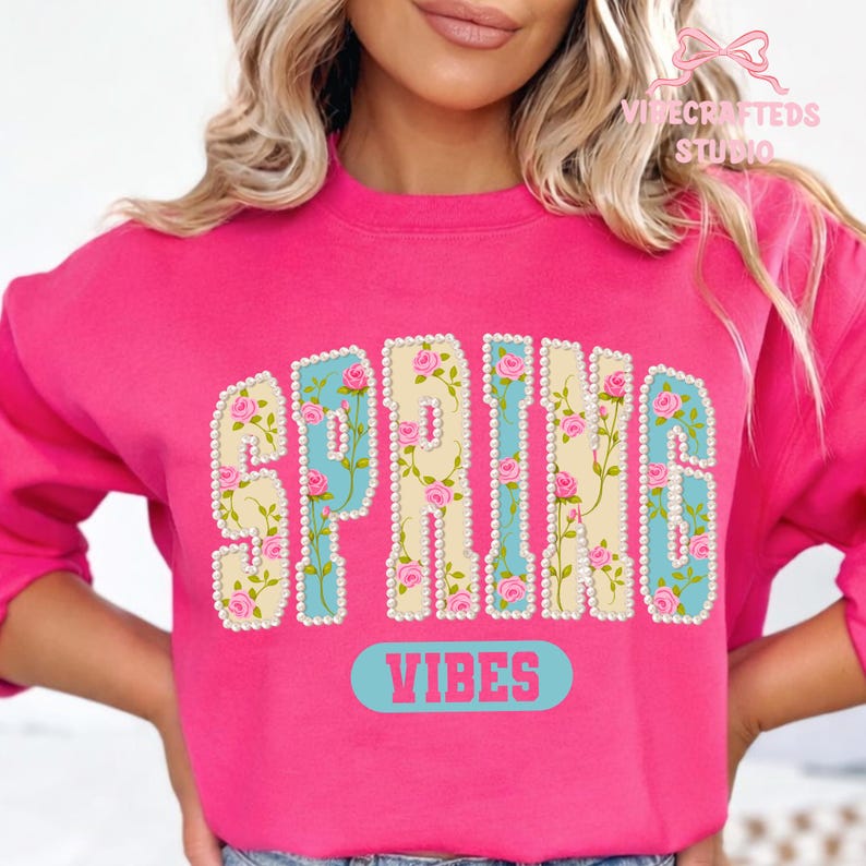 May include: A bright pink sweatshirt with the word "SPRING" in a floral design. The letters are outlined with white beads, and the background is light blue and yellow. Below the word "SPRING" is the word "VIBES" in a light blue oval.