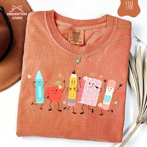 Puede incluir: Una camiseta de manga corta color terracota con un estampado gr&aacute;fico de cinco &uacute;tiles escolares antropom&oacute;rficos: un cray&oacute;n azul, una manzana roja, un l&aacute;piz amarillo, un cuaderno rosa y un pegamento en barra, todos con caras sonrientes.