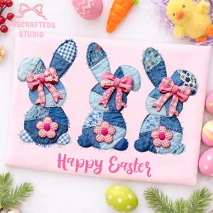 May include: A pink Easter-themed shirt featuring three denim bunny appliqu&eacute;s with pink bows and flower accents. The words "Happy Easter" are printed in pink script below the bunnies. Decorative Easter eggs and a toy chick are in the background.