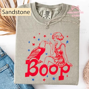 May include: Sandstone-coloured t-shirt with a red and blue graphic. The graphic features an eagle, a woman, and the word "Boop". The shirt has a crew neck and short sleeves. A silver necklace with a pendant is draped on the shirt.