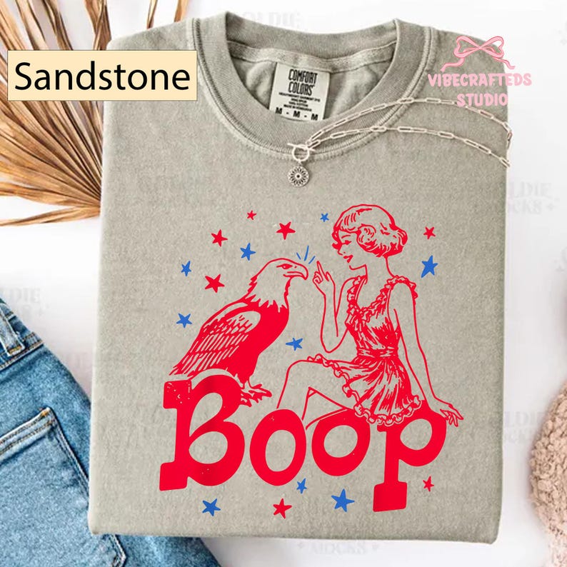 May include: Sandstone-coloured t-shirt with a red and blue graphic. The graphic features an eagle, a woman, and the word "Boop". The shirt has a crew neck and short sleeves. A silver necklace with a pendant is draped on the shirt.