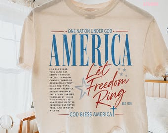 Puede incluir: Camiseta de color marfil con la palabra "AMERICA" en azul y "Let Freedom Ring" en escritura roja. La camiseta también presenta el texto "ONE NATION UNDER GOD" y "GOD BLESS AMERICA". El diseño incluye estrellas y el año "EST. 1776".