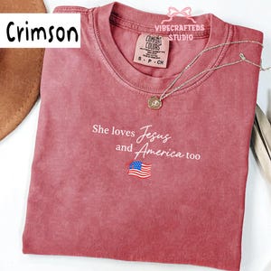 Könnte beinhalten: Karmesinrotes T-Shirt mit dem Text "She loves Jesus and America too" und einer amerikanischen Flaggen-Grafik. Das Shirt hat einen Rundhalsausschnitt und kurze Ärmel. Eine silberne Halskette mit Anhänger liegt über dem Shirt.