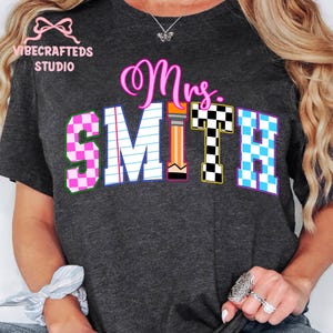 May include: Dark gray t-shirt with the text "Mrs. SMITH" in a colorful design. The "S" and "H" are checkered, the "M" is lined like notebook paper, and the "T" is a pencil. The shirt has the "VIBECRAFTED STUDIO" logo.