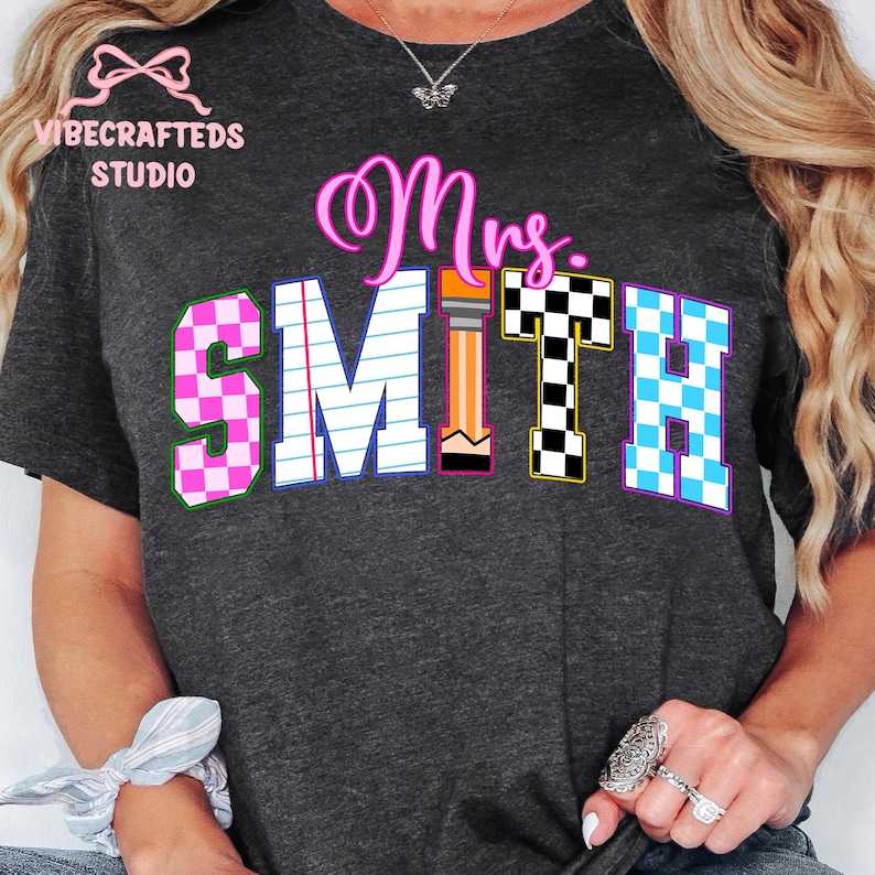 May include: Dark gray t-shirt with the text "Mrs. SMITH" in a colorful design. The "S" and "H" are checkered, the "M" is lined like notebook paper, and the "T" is a pencil. The shirt has the "VIBECRAFTED STUDIO" logo.