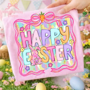 May include: A light pink t-shirt with a colorful "Happy Easter" graphic. The design features a bunny, flowers, and a carrot bow. The text is in pastel colors with a glittery background, perfect for Easter.
