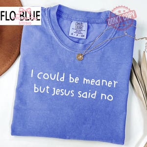May include: A blue Comfort Colors t-shirt with the text "I could be meaner but Jesus said no" in white. A gold-colored necklace with a small pendant rests on the shirt. The shirt is folded.
