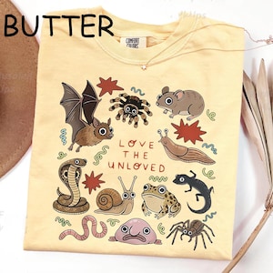 May include: Pale yellow t-shirt with a graphic of assorted animals, such as a bat, spider, and snake, and the words "LOVE THE UNLOVED." The shirt has a crew neck and short sleeves.