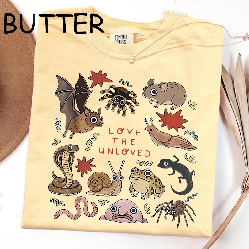 May include: Pale yellow t-shirt with a graphic of assorted animals, such as a bat, spider, and snake, and the words "LOVE THE UNLOVED." The shirt has a crew neck and short sleeves.