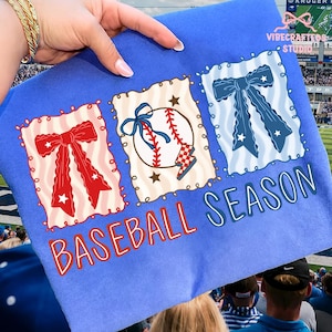 Puede incluir: Una sudadera azul de temporada de béisbol con tres cuadrados decorativos. Los cuadrados contienen un lazo rojo, una pelota de béisbol con un lazo azul y un lazo azul. Las palabras "BASEBALL SEASON" están impresas en rojo y azul.