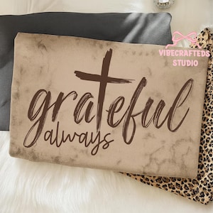 May include: A rectangular, tan-colored pillow with the words "grateful always" and a cross in brown script. The pillow is on a white surface with a dark gray pillow and a leopard print fabric in the background.