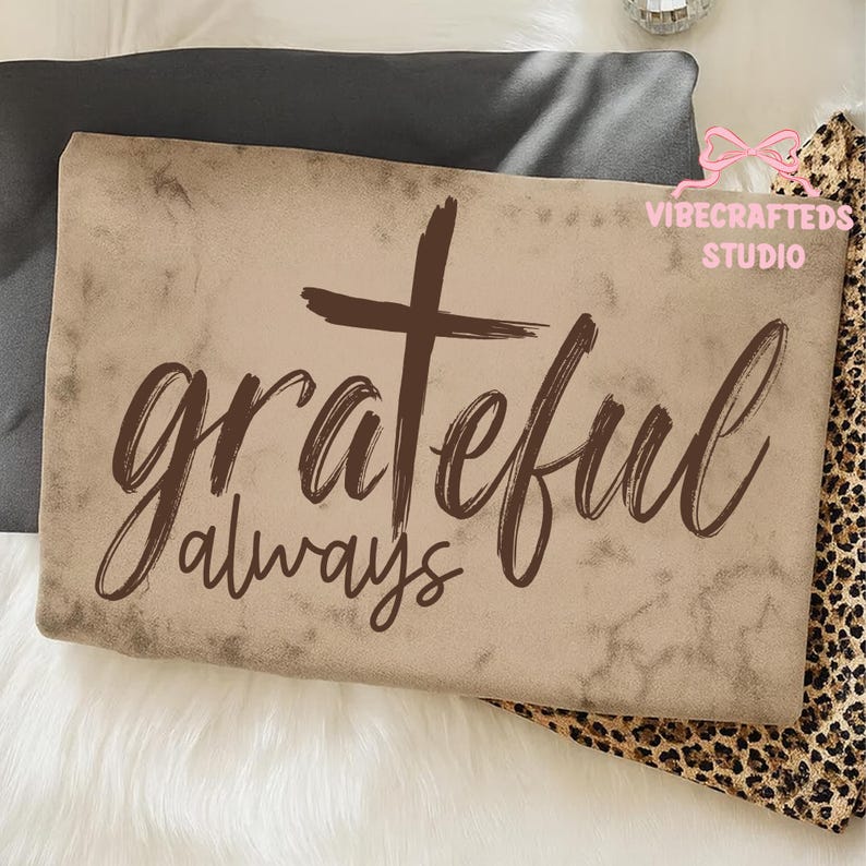 May include: A rectangular, tan-colored pillow with the words "grateful always" and a cross in brown script. The pillow is on a white surface with a dark gray pillow and a leopard print fabric in the background.