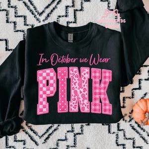 May include: A black long-sleeve sweatshirt featuring the pink text In October we Wear PINK. The letters are filled with various pink patterns including checkers, polka dots, leopard print with ribbons, and plaid, all set against a black fabric.