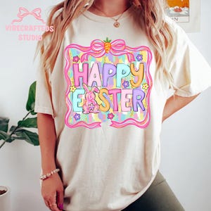 May include: Cream-colored t-shirt featuring a colorful "Happy Easter" graphic. The design includes a bunny, flowers, and a carrot, all within a decorative frame. The shirt has short sleeves and a relaxed fit, perfect for Easter celebrations.