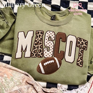 May include: A military green crewneck sweatshirt featuring the word MASCOT in large, textured applique letters. The letters alternate between cream, brown, and leopard print patterns. A brown football graphic with white laces sits below the text.