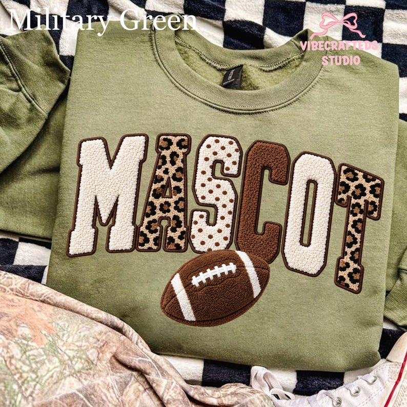 May include: A military green crewneck sweatshirt featuring the word MASCOT in large, textured applique letters. The letters alternate between cream, brown, and leopard print patterns. A brown football graphic with white laces sits below the text.
