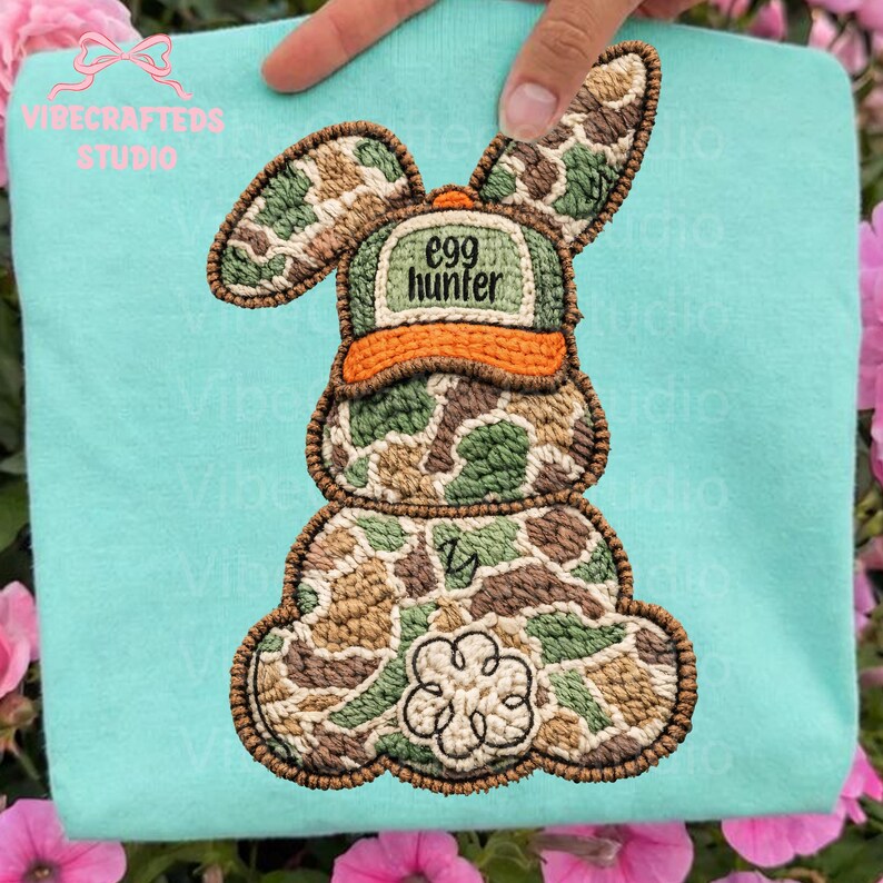 May include: An embroidered bunny design with a camouflage pattern, wearing an orange-brimmed hat that reads "egg hunter." The bunny is on a light blue background, with a flower design on its lower body. The design is likely for a shirt or other apparel.