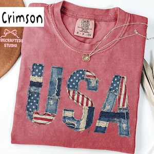 Op de afbeelding: Karmijnrood T-shirt met het woord "USA" in een patchwork-ontwerp met de Amerikaanse vlag. Een zilveren ketting met een hanger ligt op het shirt. Het shirt heeft een Comfort Colors label.