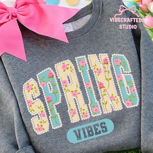 May include: A gray sweatshirt with the word "SPRING" in a floral design, with pink roses and a light blue background. The word "VIBES" is below the word "SPRING". A large pink bow is in the upper left corner.