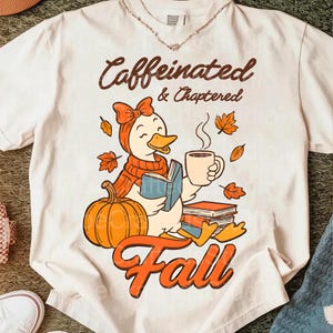 Puede incluir: Camiseta blanca con un diseño de otoño. El diseño presenta un pato de dibujos animados con bufanda y lazo, sosteniendo un libro y café, con una calabaza y las palabras "Caffeinated & Chaptered Fall".