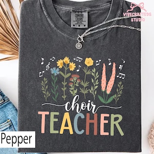 May include: A dark gray t-shirt with a floral design and musical notes. The shirt features the words "choir TEACHER" in colorful letters. A silver necklace with a pendant is on the shirt. The shirt is labeled "Pepper".
