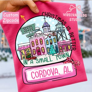 May include: A pink t-shirt featuring a snow globe design with the text "Christmas is Better in a Small Town." The snow globe includes illustrations of buildings, a Christmas tree, and the town name "Cordova, AL." The zip code "35550" is also displayed.