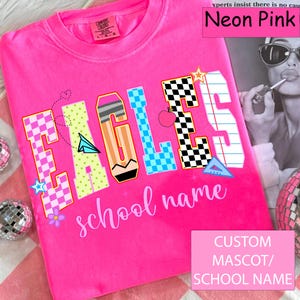 Puede incluir: Camiseta rosa neón con la palabra "EAGLES" en un diseño colorido con temática escolar. La camiseta también presenta el texto "school name". La camiseta es de la marca Comfort Colors.