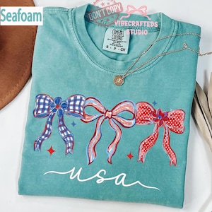 May include: Seafoam green t-shirt with a patriotic bow design. The shirt features three bows in red, white, and blue, with the word "usa" in a cursive font. A silver necklace with a pendant is visible.