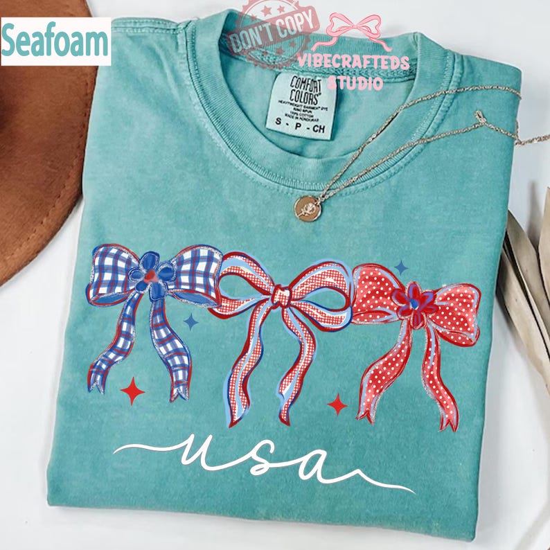 May include: Seafoam green t-shirt with a patriotic bow design. The shirt features three bows in red, white, and blue, with the word "usa" in a cursive font. A silver necklace with a pendant is visible.