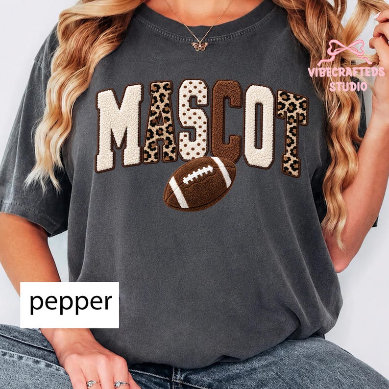 May include: A charcoal gray short-sleeve t-shirt featuring the word MASCOT in large, textured varsity-style letters. The letters alternate between cream, brown, and leopard print patterns. A brown football graphic is centered below the text.