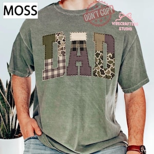 Pode incluir: Camiseta verde musgo com a palavra "DAD" em letras de patchwork. As letras apresentam vários padrões, incluindo camuflagem e xadrez. O canto superior esquerdo tem a palavra "MOSS" numa caixa branca.