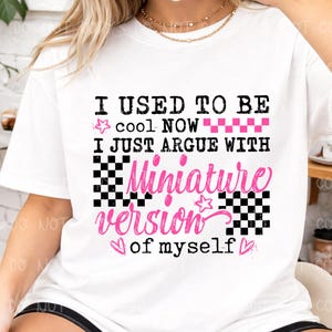 以下が含まれることがあります： 「I USED TO BE cool NOW I JUST ARGUE WITH Miniature version of myself」の文字が黒とピンクでプリントされた白いTシャツ。星、ハート、市松模様のデザインが含まれています。