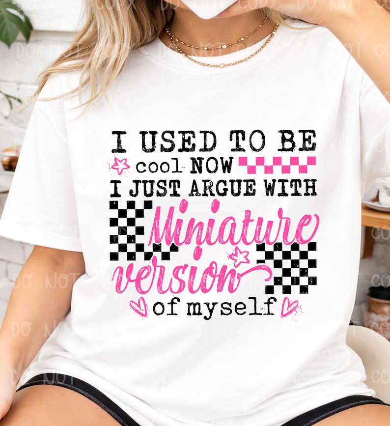 以下が含まれることがあります： 「I USED TO BE cool NOW I JUST ARGUE WITH Miniature version of myself」の文字が黒とピンクでプリントされた白いTシャツ。星、ハート、市松模様のデザインが含まれています。
