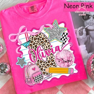Pode incluir: Uma t-shirt rosa néon com uma impressão gráfica com o nome Olivia em letra cursiva rosa. O design inclui uma forma com padrão de leopardo, uma estrela brilhante, uma bola de discoteca rosa, uma mochila rosa com xadrez, um lápis e um laço.