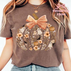 May include: A brown t-shirt featuring a pumpkin design made of flowers, ghosts, and pumpkins. The pumpkin has a large orange bow on top. The shirt has the text "VIBECRAFTED STUDIO" in the upper right corner.