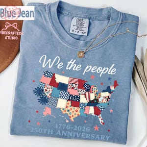 May include: A pale blue t-shirt featuring the phrase "We the people" and a patchwork map of the United States. The shirt also displays the text "1776-2026 250th Anniversary". A gold necklace is draped over the shirt.