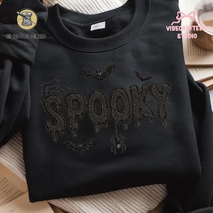 May include: A black crewneck sweatshirt featuring black embroidery of the word SPOOKY in a dripping font. The design includes embroidered details of a spider web, three bats, and a spider hanging from the letter K on the front of the garment.