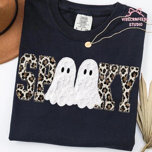 May include: A black short-sleeved t-shirt featuring the word SPOOKY in block letters with a leopard print pattern. The two O's are replaced by white lace ghosts. The shirt has a Comfort Colors label at the neck and a gold chain necklace.