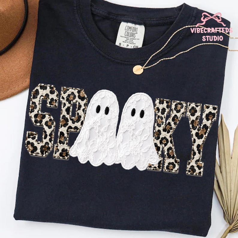 May include: A black short-sleeved t-shirt featuring the word SPOOKY in block letters with a leopard print pattern. The two O's are replaced by white lace ghosts. The shirt has a Comfort Colors label at the neck and a gold chain necklace.
