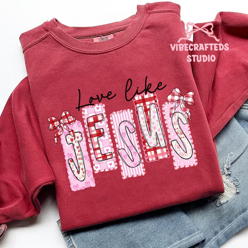May include: A maroon sweatshirt with the words "Love like Jesus" in black script. The word "JESUS" is in a patchwork design with pink and red patterns, hearts, and bows. The sweatshirt is folded over denim shorts.
