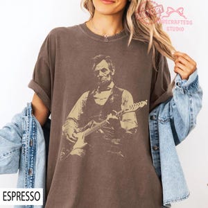 May include: Espresso-colored t-shirt featuring a sepia-toned graphic of a man playing an electric guitar. The graphic appears to be Abraham Lincoln. The shirt has rolled-up sleeves and the word "ESPRESSO" is printed at the bottom left.