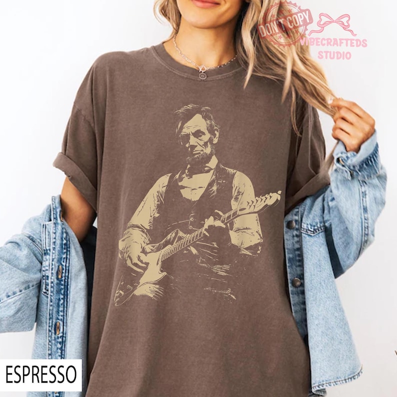 May include: Espresso-colored t-shirt featuring a sepia-toned graphic of a man playing an electric guitar. The graphic appears to be Abraham Lincoln. The shirt has rolled-up sleeves and the word "ESPRESSO" is printed at the bottom left.