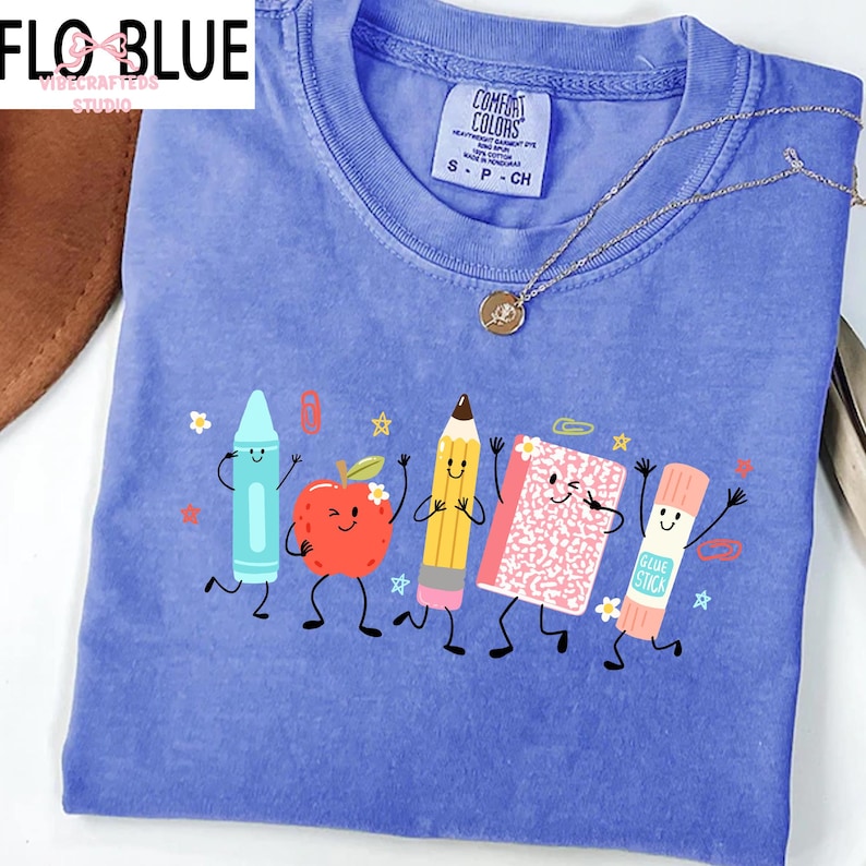 Puede incluir: Una camiseta de color azul b&iacute;garo con cuello redondo que presenta un estampado gr&aacute;fico de cinco &uacute;tiles escolares antropom&oacute;rficos, incluyendo un cray&oacute;n azul claro, una manzana roja, un l&aacute;piz amarillo, un cuaderno rosa y un pegamento.