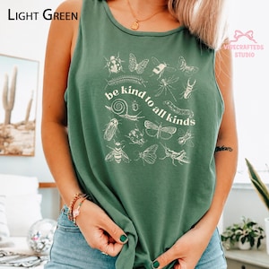May include: A light green sleeveless tank top featuring a white graphic print of various insects and the text be kind to all kinds. The design includes illustrations of a snail, butterfly, bee, and other small creatures arranged in a central cluster.