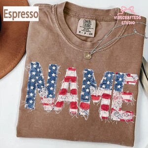 May include: Espresso-coloured t-shirt featuring the word "NAME" in a distressed American flag design. The shirt has a crew neck and short sleeves. The tag reads "Comfort Colors". A silver necklace with a pendant is visible.