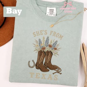 May include: A light blue t-shirt with the text "She's From Texas" and a graphic of brown cowboy boots, flowers, and a horseshoe. The shirt has a crew neck and short sleeves.